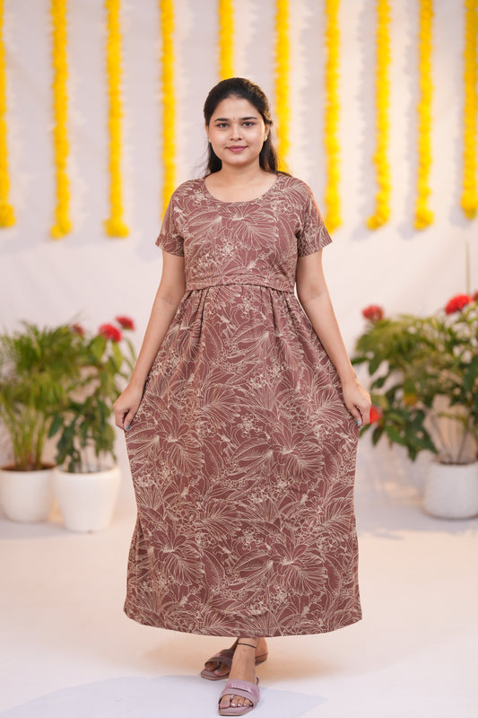 A woman modeling a brown and beige tropical leaf print maxi dress with short sleeves and a gathered waist, standing in front of a decorated white wall with yellow garlands and potted plants.